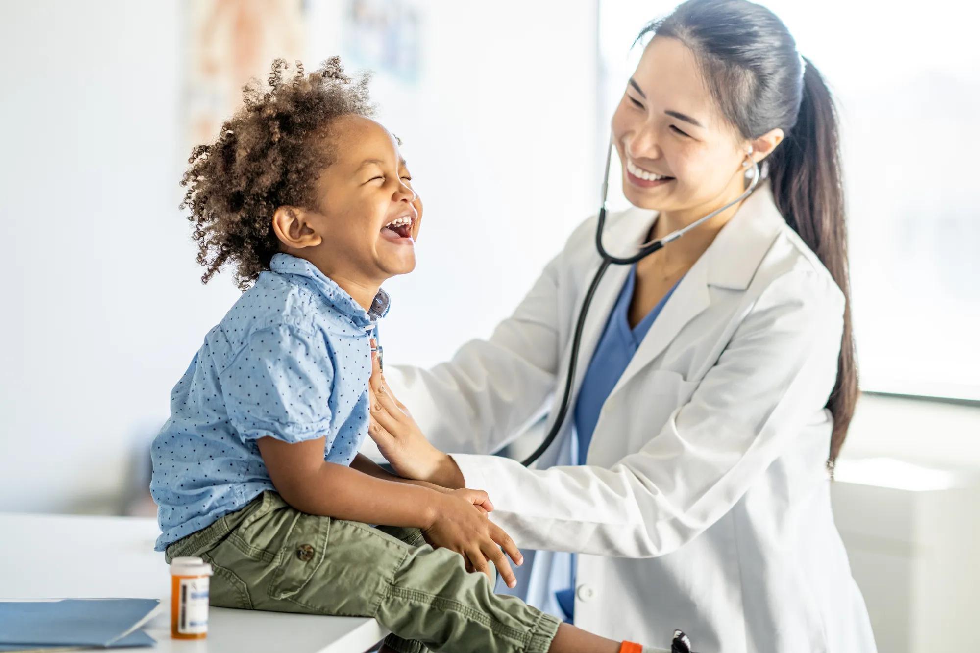 A sweet young mixed race boy, sits up on an exam able with a big smile on his face, during a routine check-up.  His female doctor of Asian decent is leaning in with her stethoscope to listen to his heart.