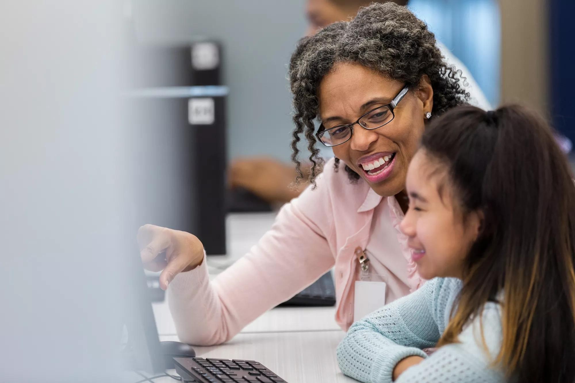 African American female school teacher points to something on computer while helping a female student during technology class.